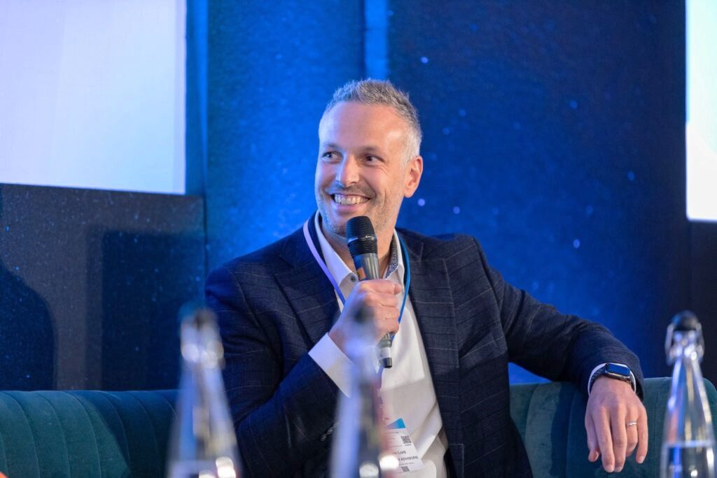 A businessman in a suit smiles while holding a microphone at a Carbon Forward panel discussion.